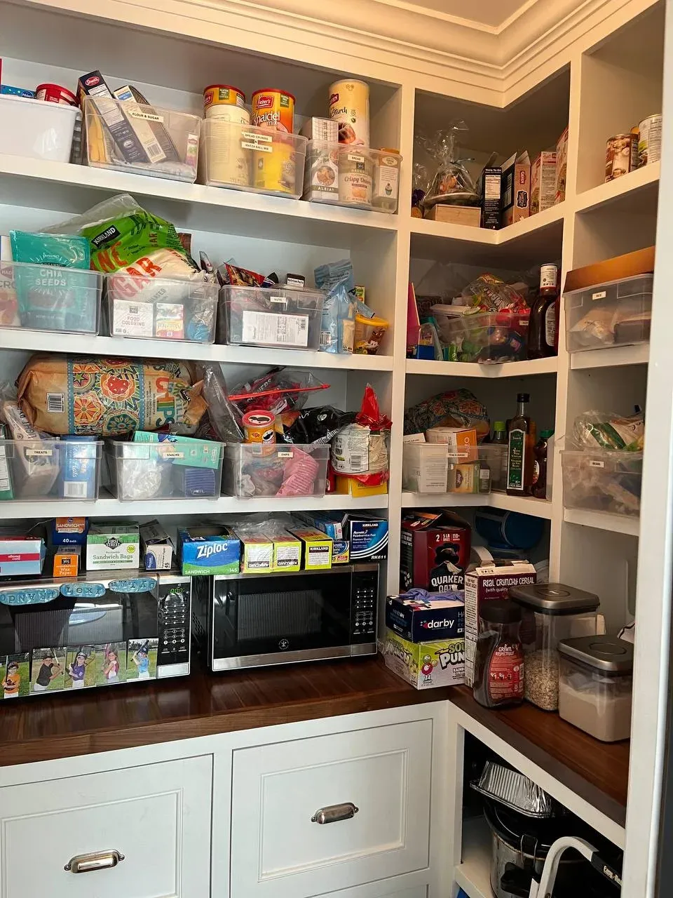 Pantry corner before, shelves packed with mismatched boxes, bags, and bins stacked on top of each other above two microwaves on the counter