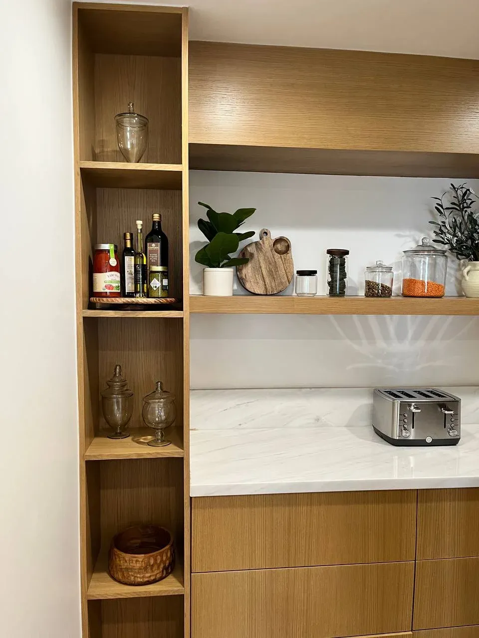 Kitchen pantry from the side, with a tall oak cubby column of oils and jars beside the floating shelf of decanted dry goods