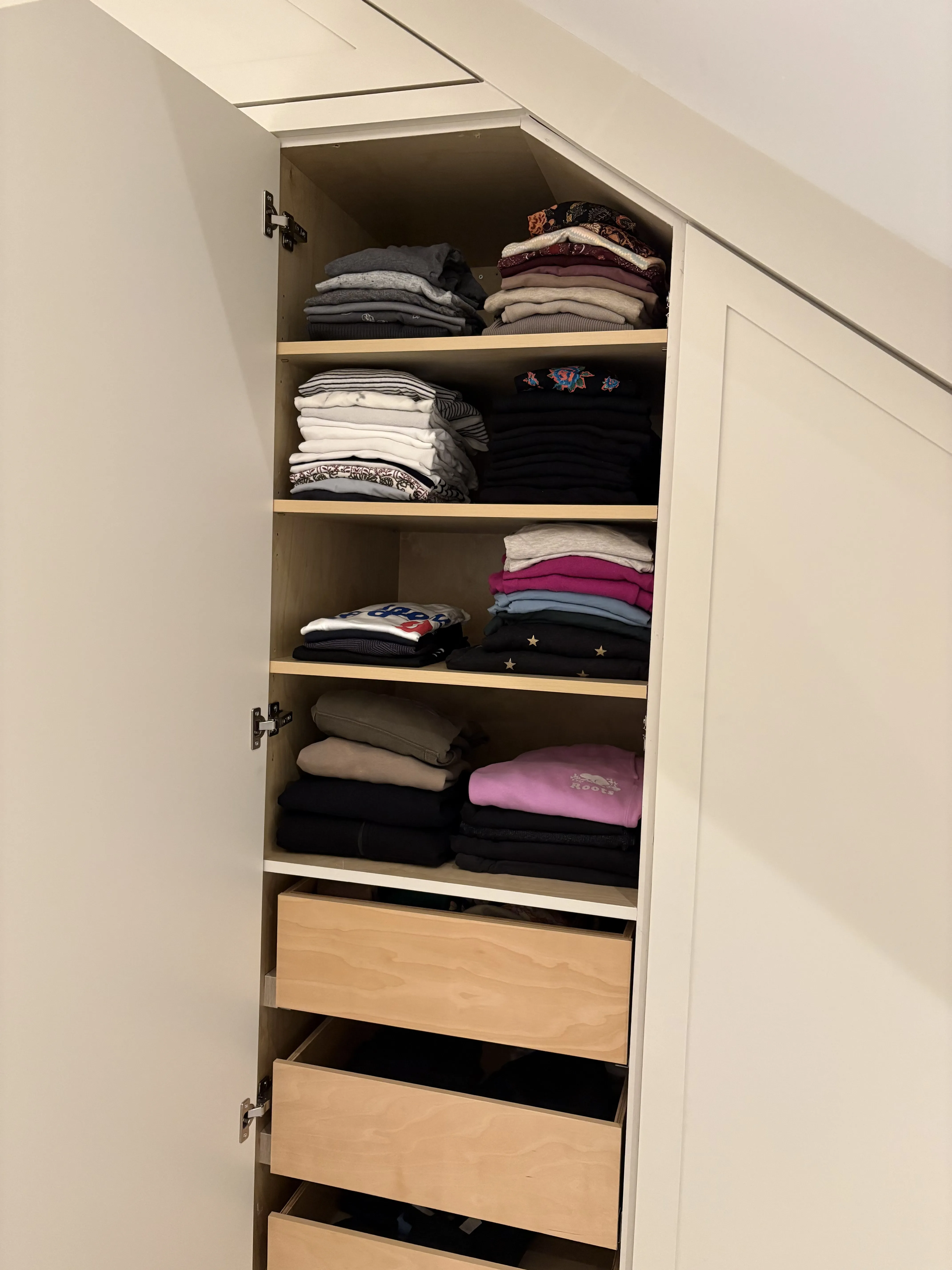 Tall closet under a sloped ceiling with four shelves of folded tees, hoodies, and sweatshirts and three soft-close drawers underneath