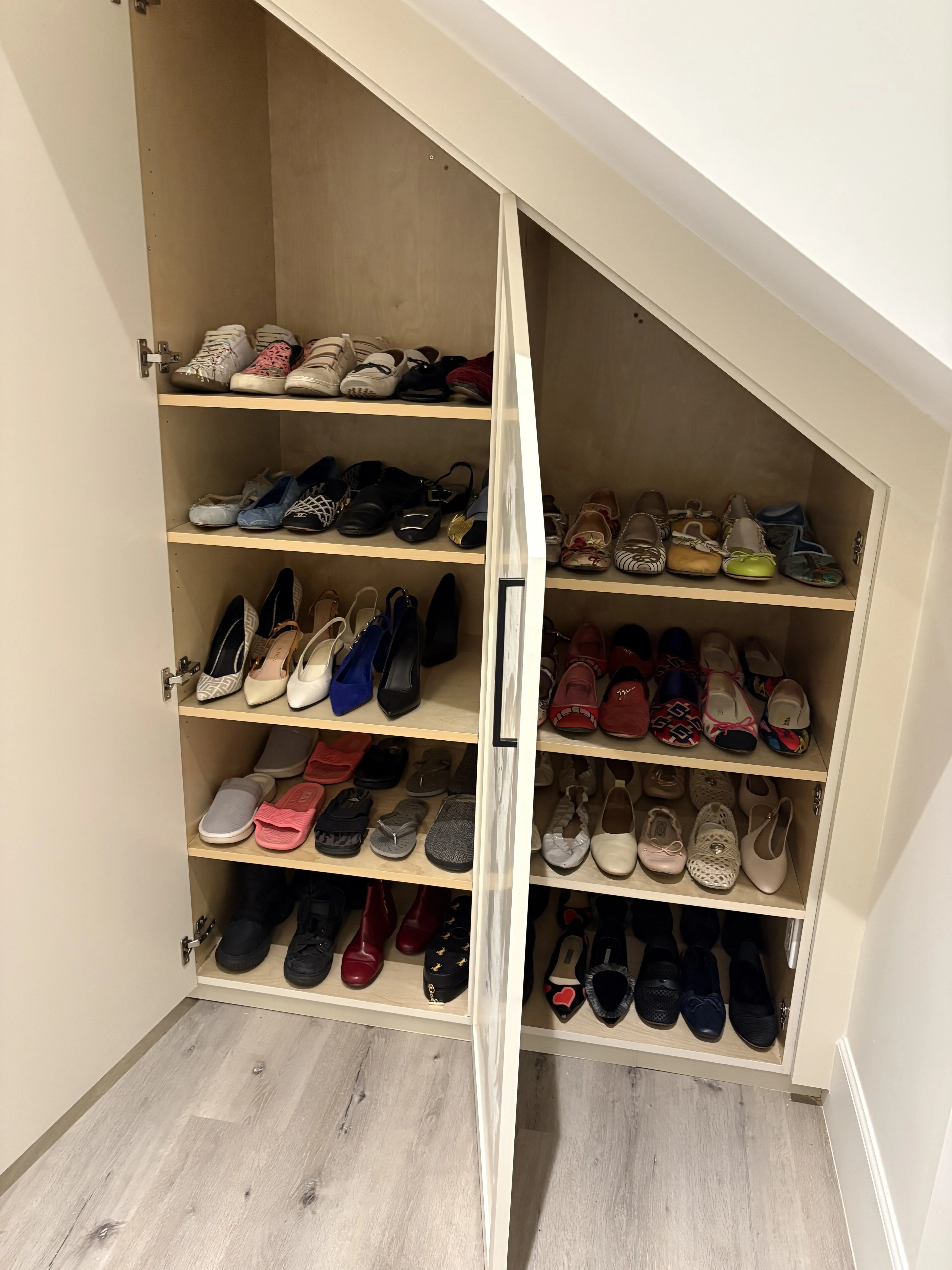 Shoe closet tucked under the staircase slope, with open shelves of sneakers, flats, heels, and boots on both sides of a central divider