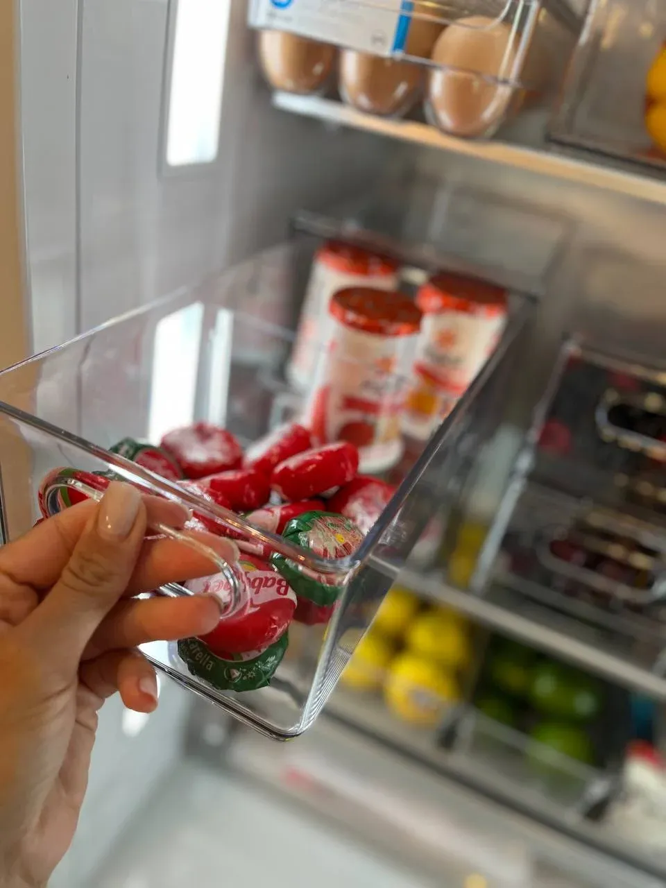 A hand holding a clear fridge bin of Babybel cheese, with eggs and citrus on the shelves behind