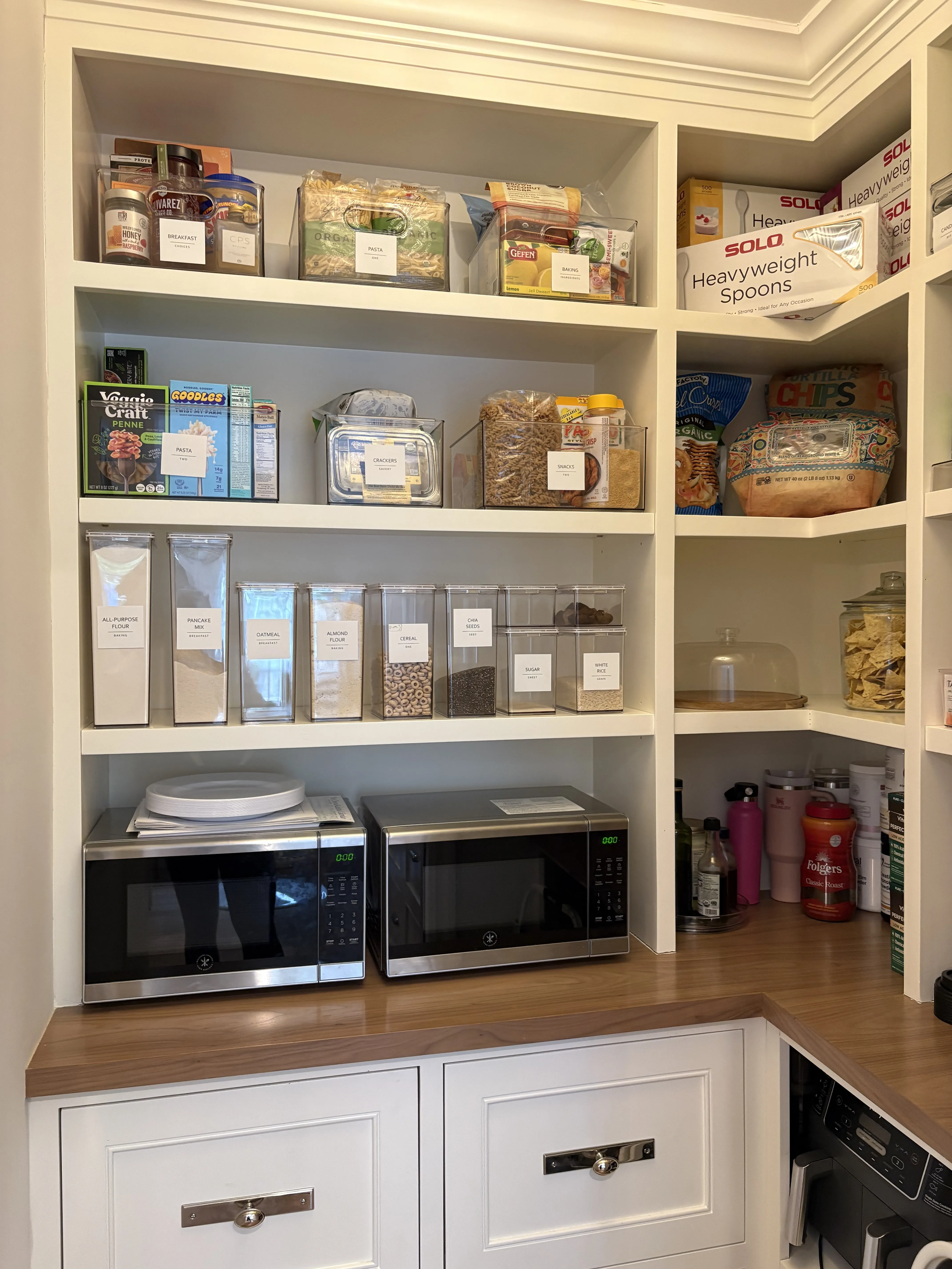 Small pantry with open white shelves holding labeled clear bins, decanted dry-goods containers in a row, and two microwaves on a butcher-block counter above white drawers