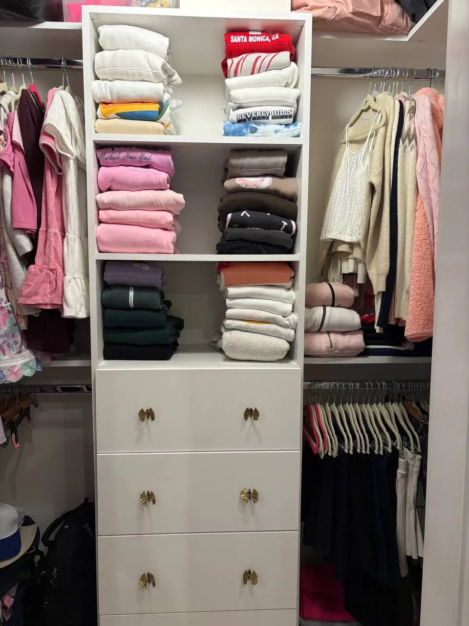 A girl's white built-in closet with open shelves of color-sorted sweatshirts, a bank of gold-knob drawers in the center, and clothes hanging on both sides