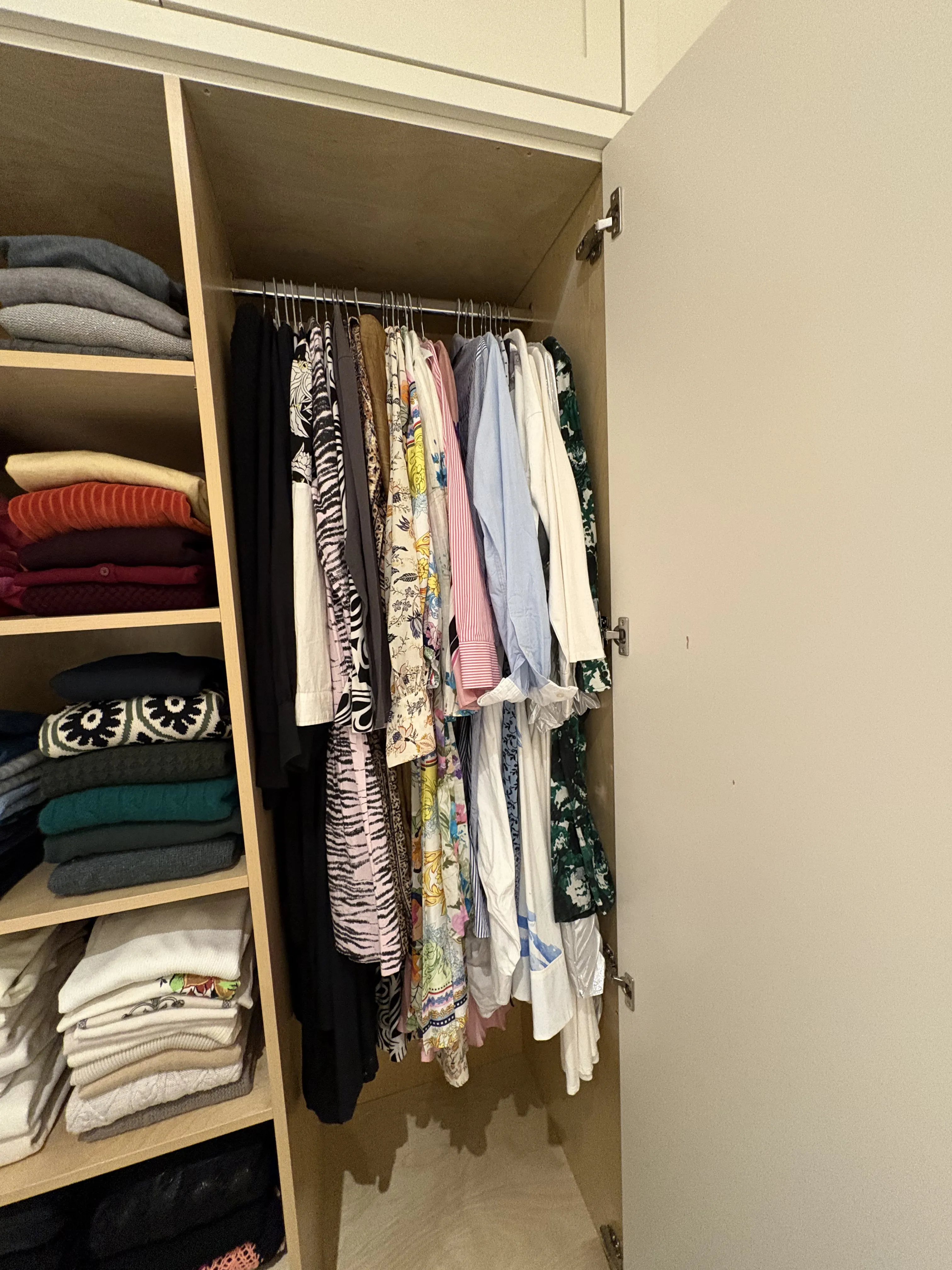 Tall closet with one side of folded sweaters and knits stacked on open shelves, and the other side hung with patterned blouses and light tops on matching hangers