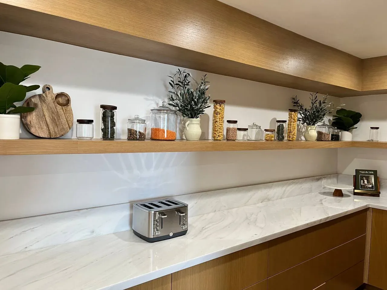 Floating oak kitchen shelf styled with decanted glass canisters of pasta and grains, olive branches in white pitchers, and cutting boards, above a marble counter