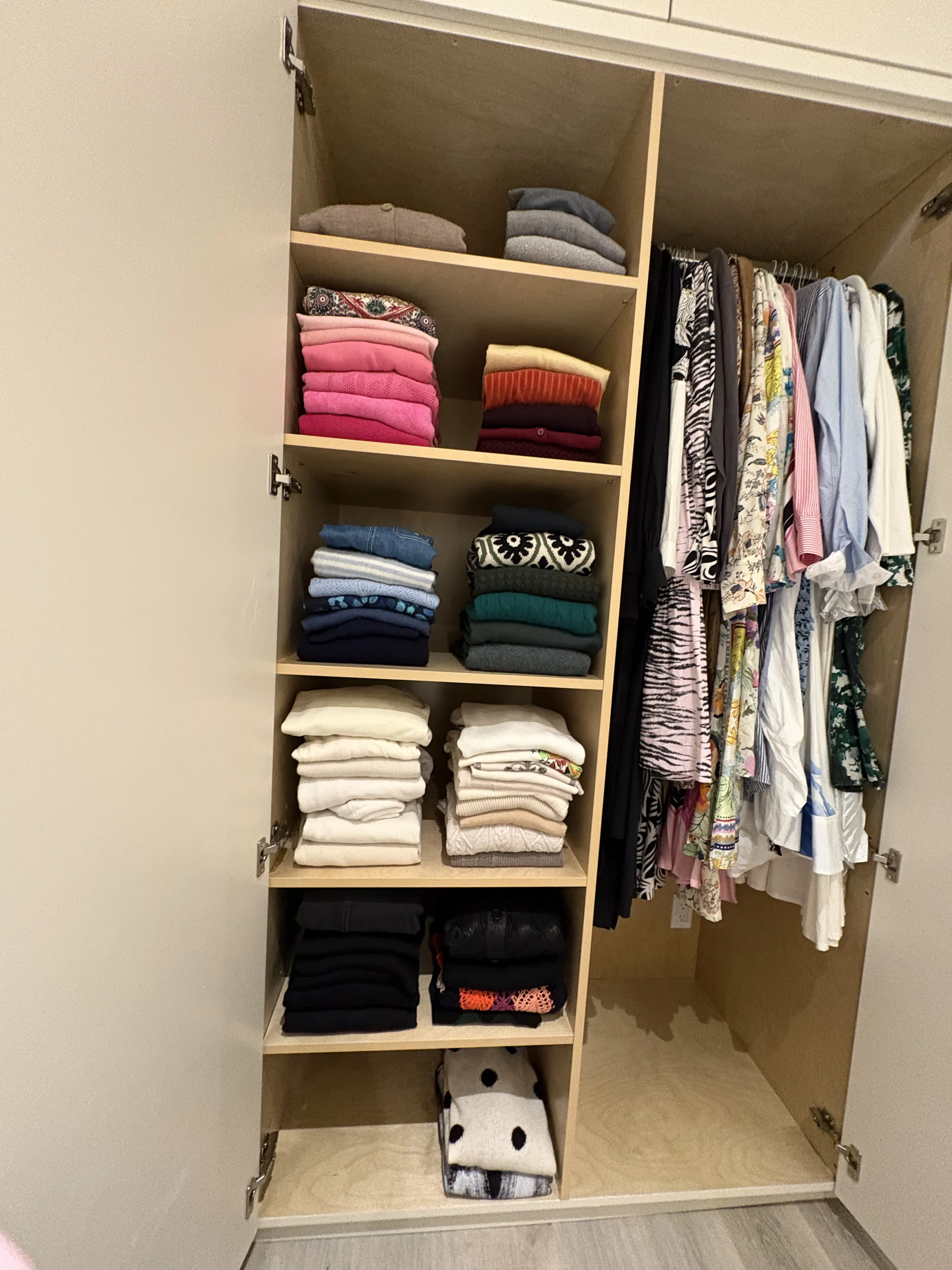 Tall closet with stacked folded sweaters in colored zones on one side, and a row of patterned blouses and light tops hung on matching hangers on the other