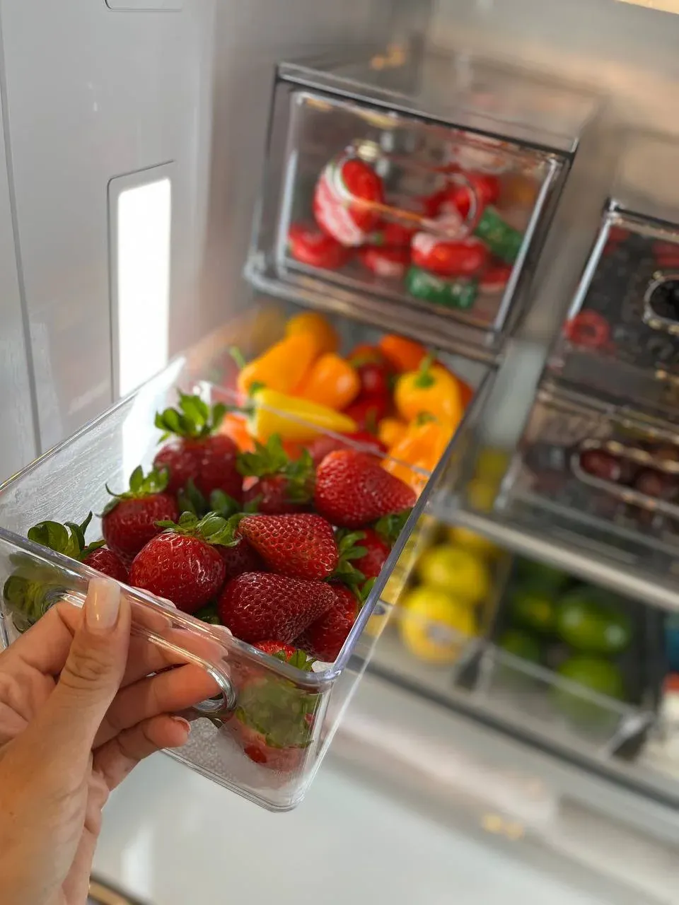 A hand lifting a clear bin of strawberries from the fridge, with peppers, lemons, and limes sorted by color in the drawers below