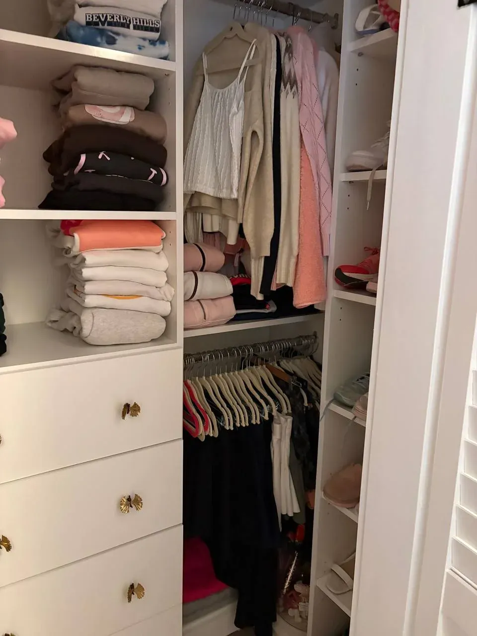 Corner of the girl's walk-in with a shelf tower of folded sweatshirts, hanging cardigans, and a column of shoes on the right