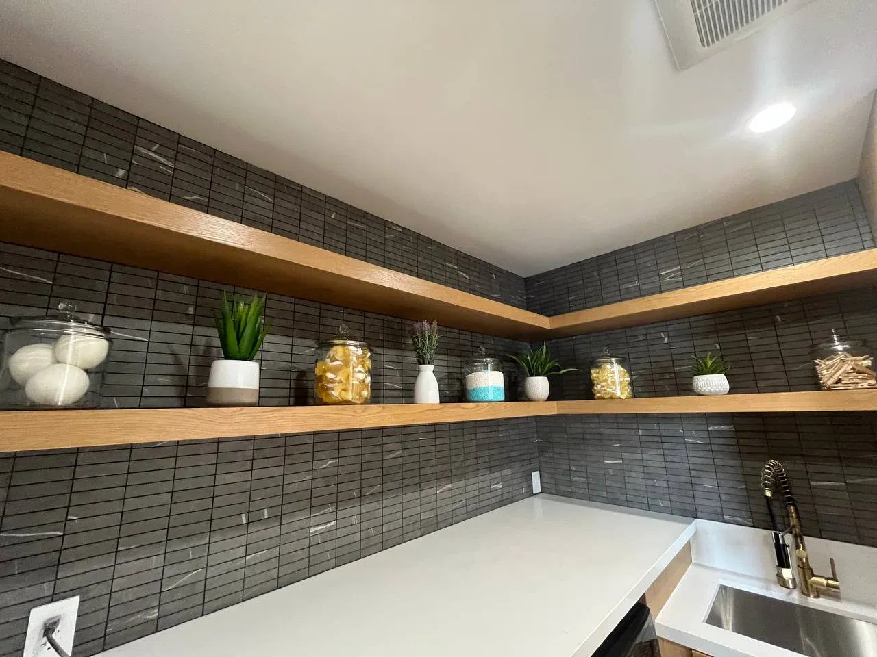 Laundry room with oak floating shelves over dark stacked tile, glass jars of dryer balls and detergent, small plants, and a brass faucet