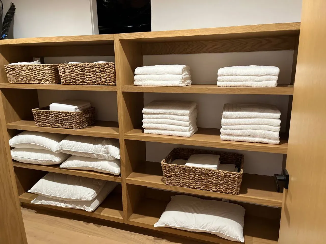 Open oak linen shelves with white towels folded in neat stacks, woven baskets, and pillows squared away on the lower shelves