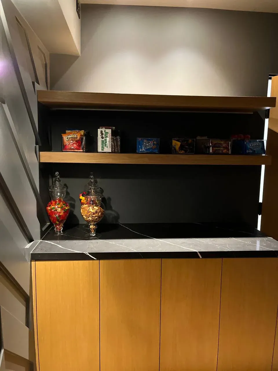 Snack station tucked under a staircase, with apothecary jars of candy and trail mix on dark marble and labeled snacks in clear bins on the oak shelf above